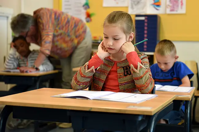 Kinder sitzen in der Schule an Tischen und eine Lehrerin steht bei einem Kind.